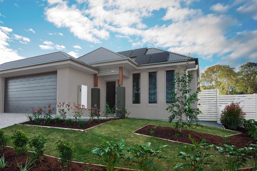 A Tesla Powerwall 3 is mounted on the exterior wall of a house, accompanied by a rooftop solar panel system. The Tesla battery stores solar power for backup energy and off-grid living.
